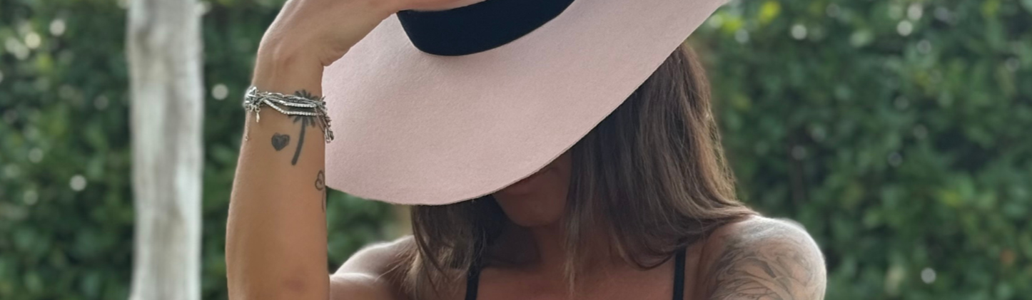 Woman with tattoos wearing a wide-brimmed hat and black swimsuit against a green background