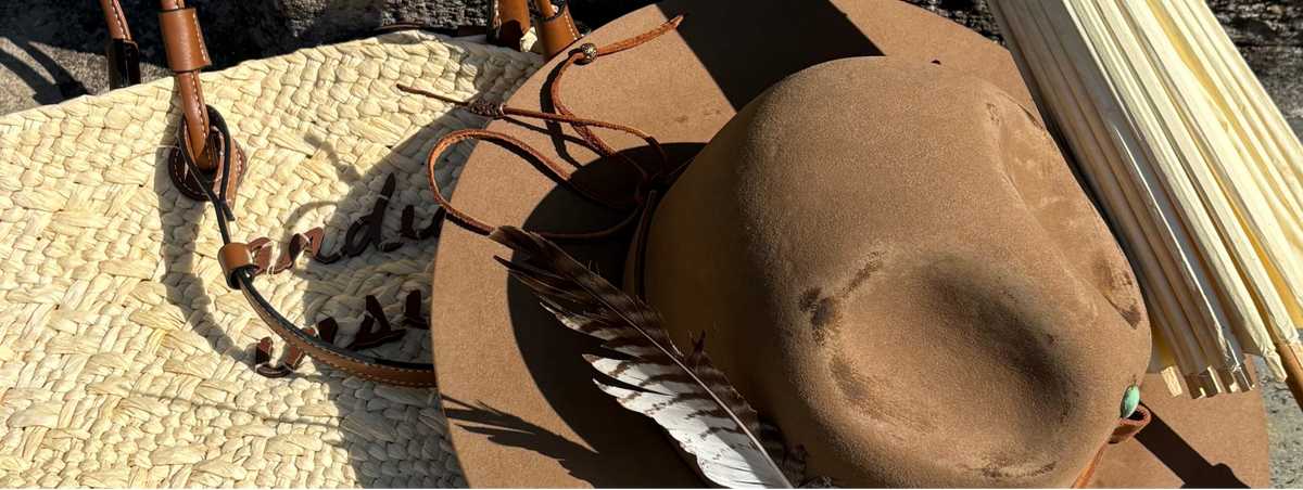 A brown felt cowboy hat with leather straps, a feather, and a woven straw hat, styled with a bundle of bamboo poles against a stone wall.