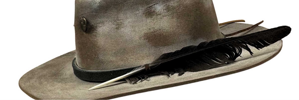 A distressed brown leather cowboy hat with a black band, a black feather, and a silver-toned metal spike, shown on a plain white background.