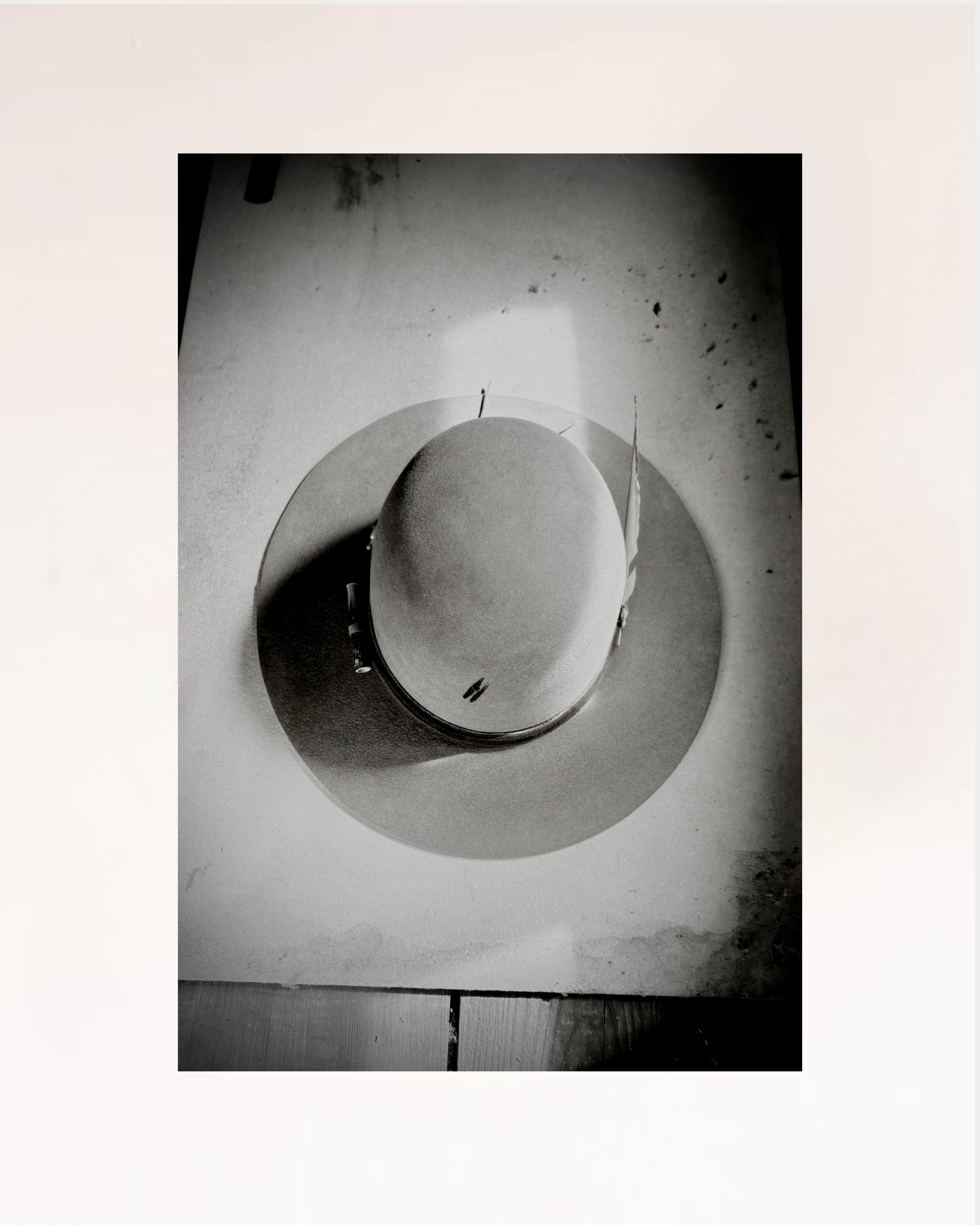 A black and white square photograph of a wide-brimmed hat resting on a circular mat against a light background, presented with a wide white border.
