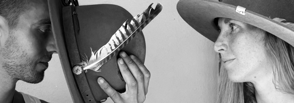 Black and white close-up of a man holding a wide-brim hat with a feather while looking at a woman wearing a matching wide-brim hat.