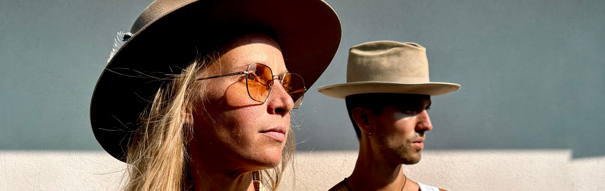 A woman in the foreground wears a wide-brimmed brown hat, round amber-tinted sunglasses, and a cream top, while a man in the background wears a beige wide-brimmed hat, a white tank top, and a gold pendant necklace against a minimalist beige and gray wall.