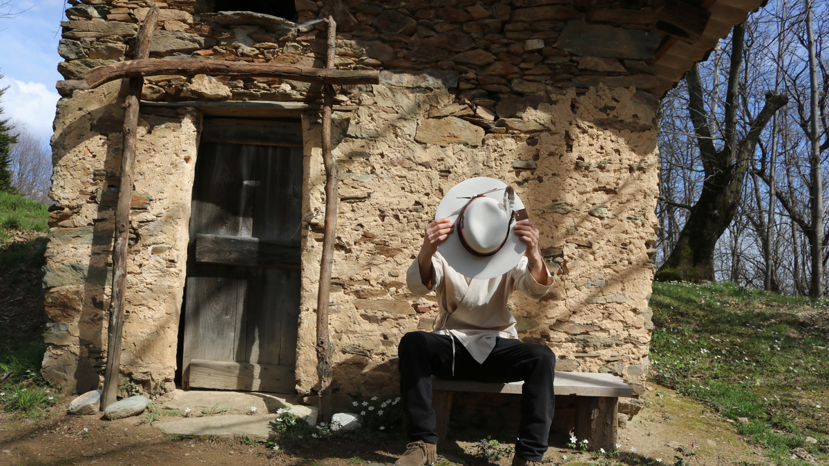Person sitting on a bench in front of a rustic stone building with bare trees in the background.